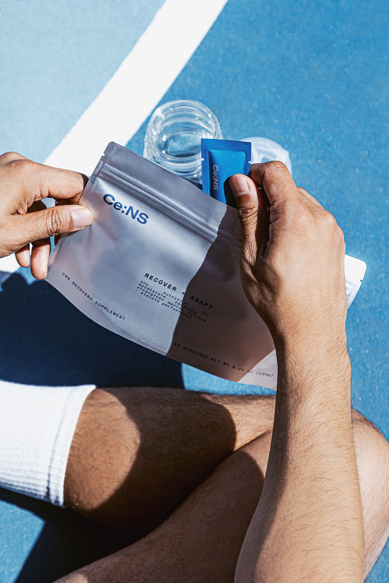 A person seated on a blue athletic court holds a pouch and packet of Ce:NS recovery supplement.