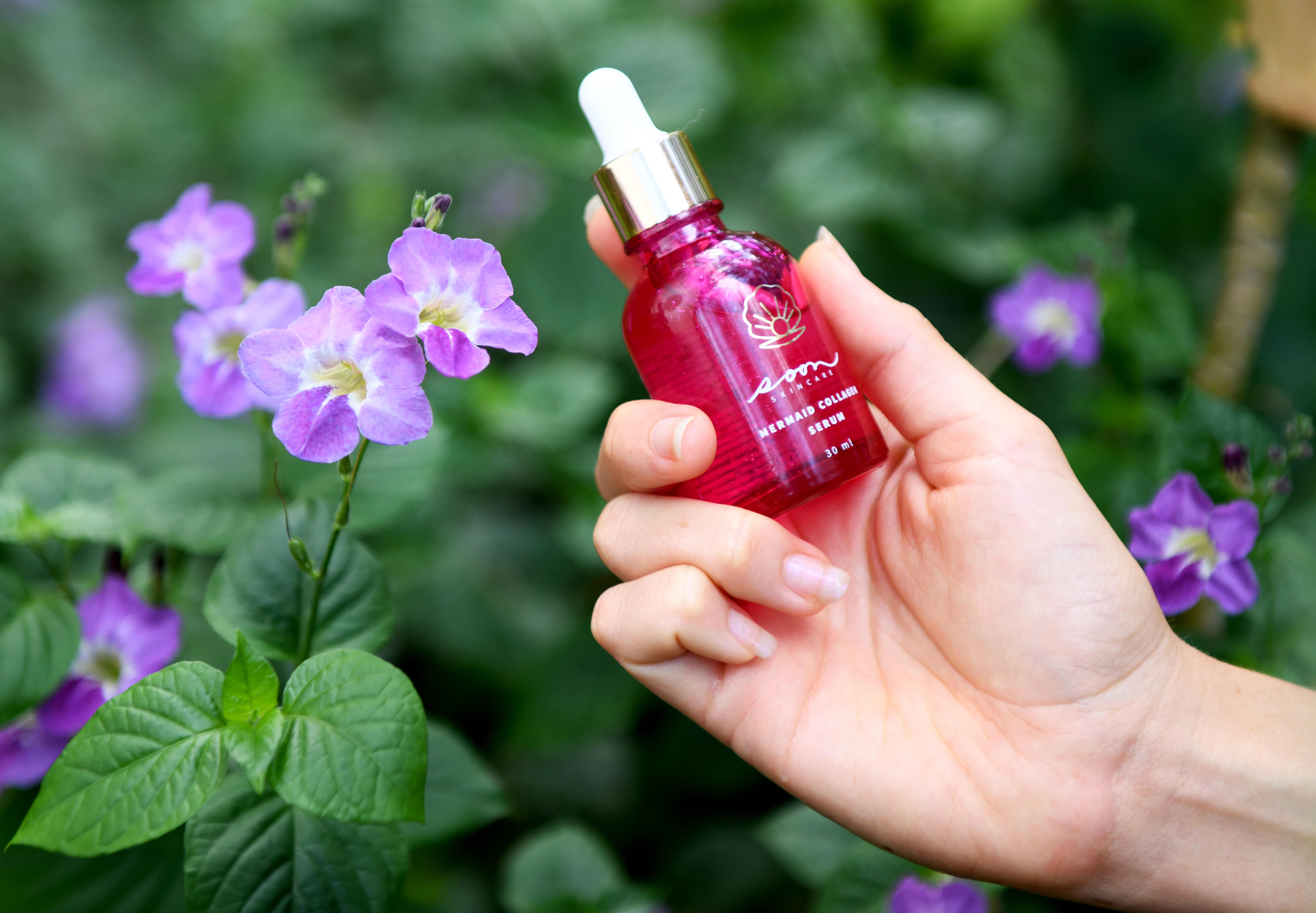 Hand holding a red serum bottle near purple flowers in a garden.