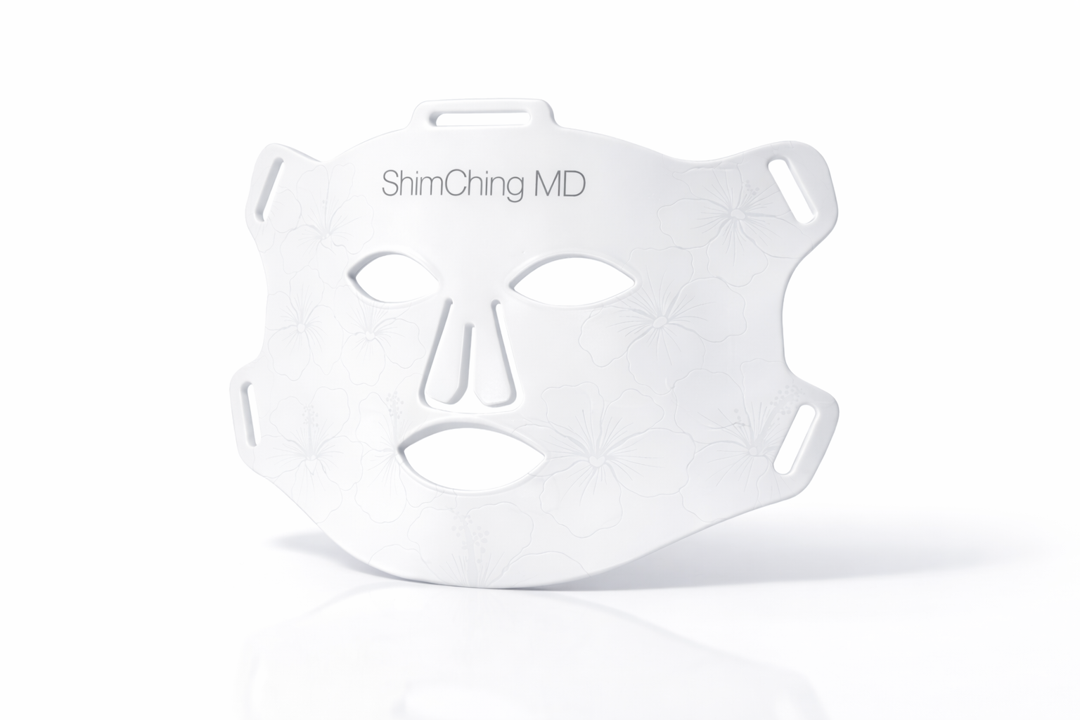 A white ShimChing MD facial mask with an embossed floral pattern, on a white reflective surface.
