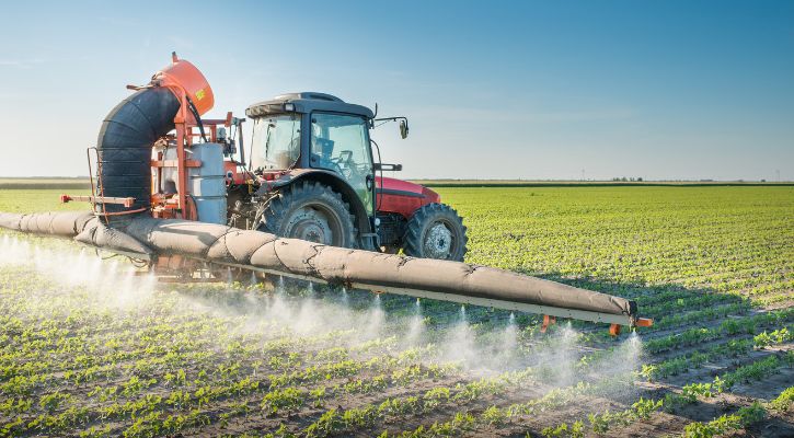 Red tractor spraying crops in a large green field under a clear blue sky.