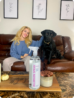 Woman sitting on a couch with a black dog, allergen spray bottle in foreground.