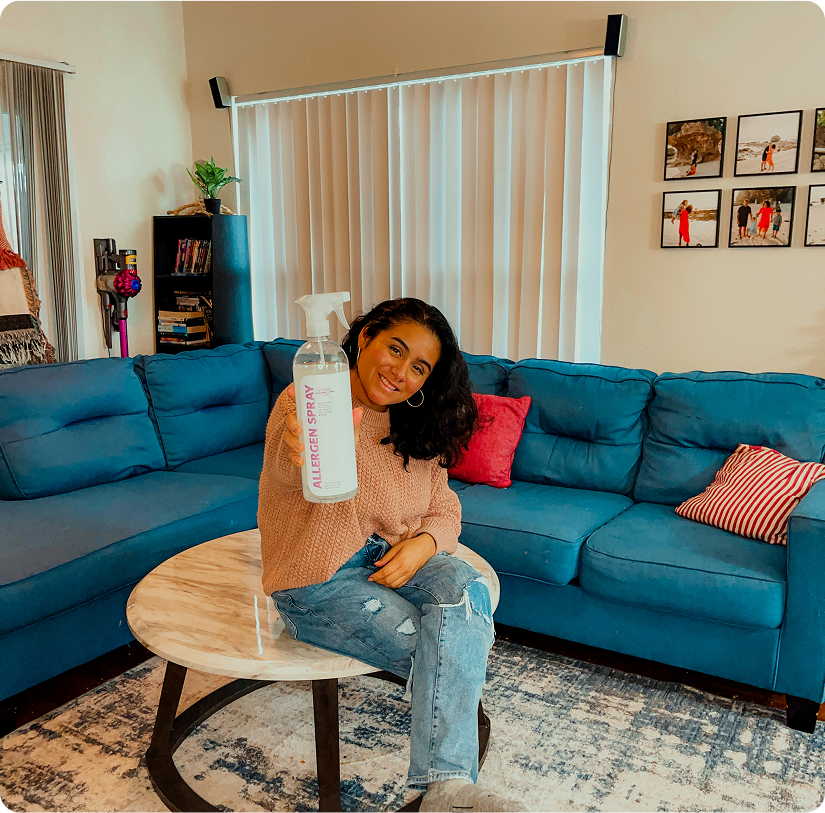 Person sitting on a couch, holding a bottle labeled 'Allergen Spray' in a living room.