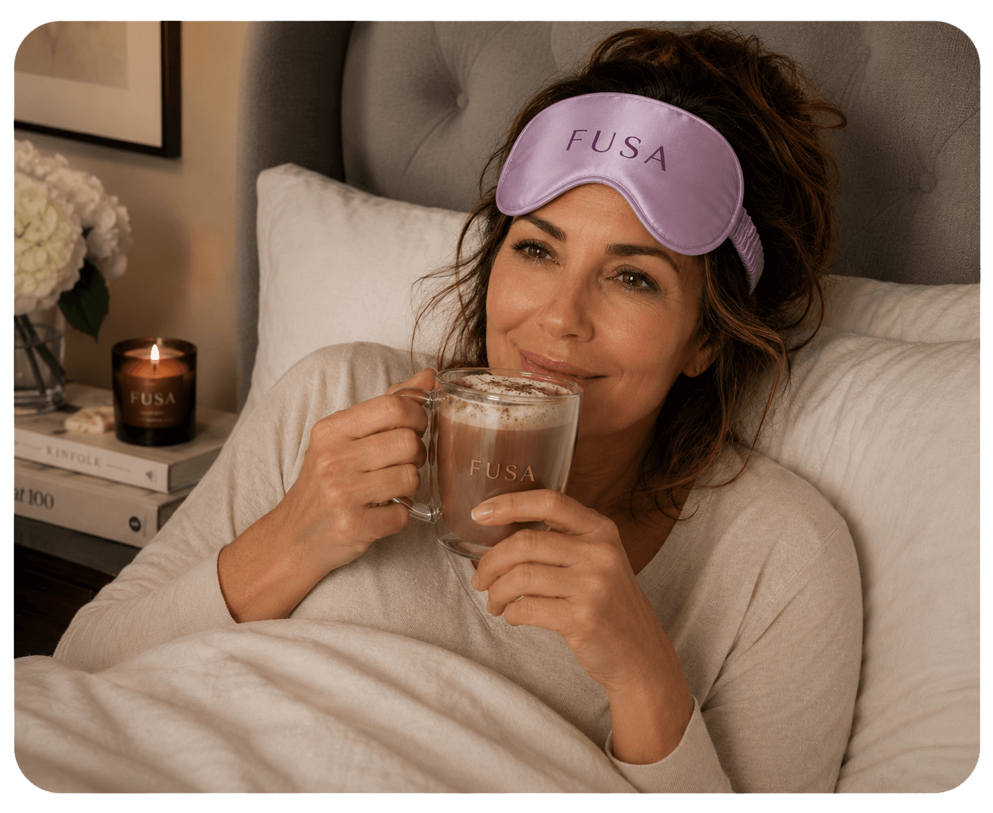 A woman relaxes in bed, holding a mug of a warm drink with a sleep mask on her head.