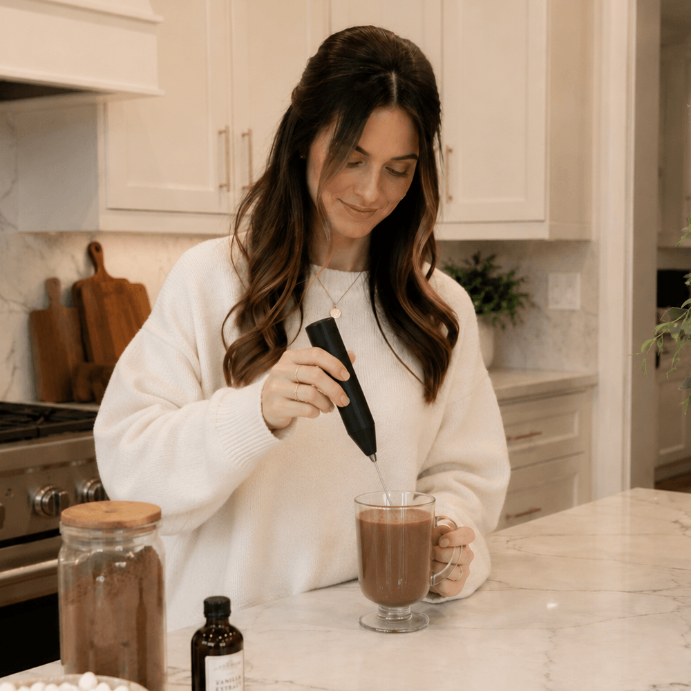 A woman in a white sweater uses a handheld frother to mix a hot beverage in a glass mug.