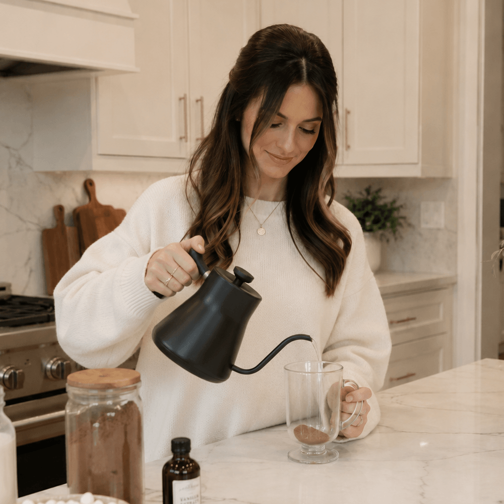 A woman in a white sweater pours hot water from a black gooseneck kettle into a clear glass mug.
