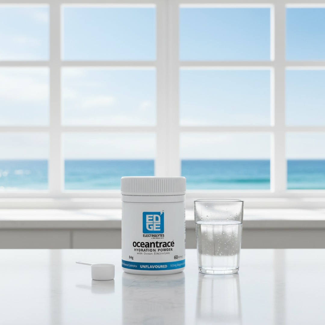 Ocean Trace hydration powder tub on a white counter with a measuring scoop and a glass of water, set in front of a large window overlooking the ocean.