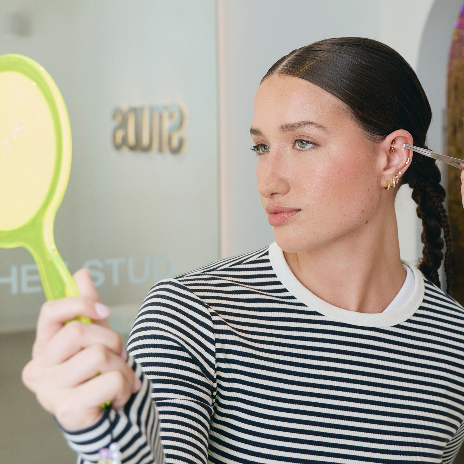 A woman holding a hand mirror getting an ear piercing consultation.