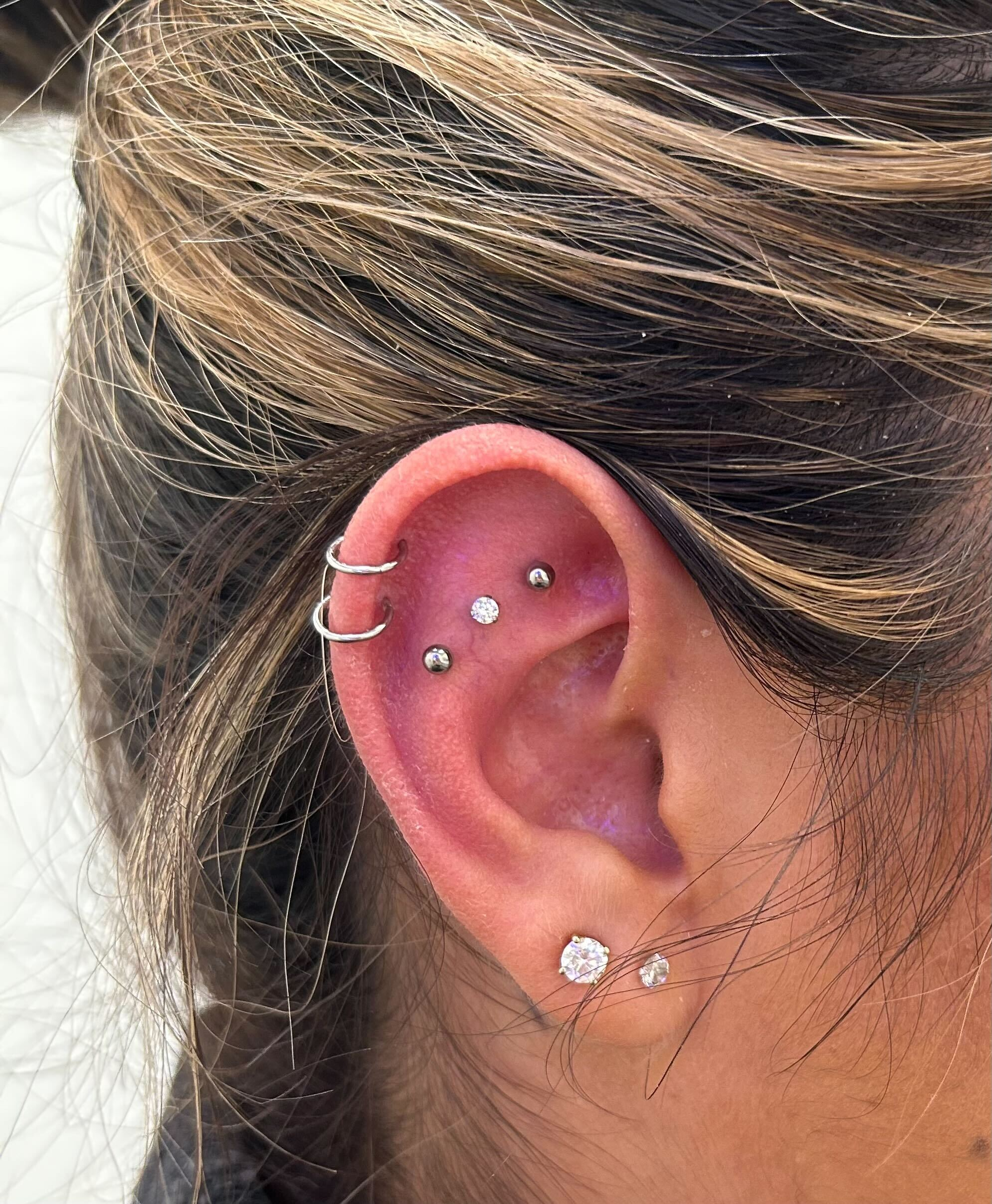 A close-up of a person's ear with multiple piercings, including silver hoops, studs, and diamond-like earrings.
