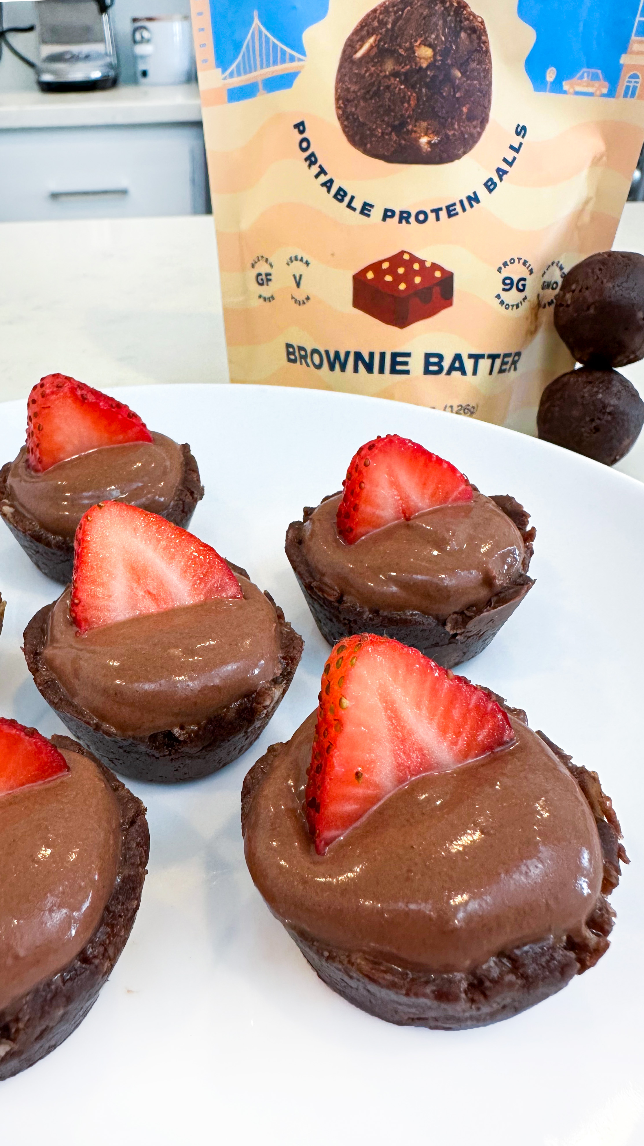 Chocolate desserts topped with strawberries on a plate, with a protein balls package in the background.