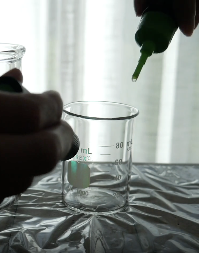 A hand uses a green dropper to put a drop of liquid into a glass beaker.