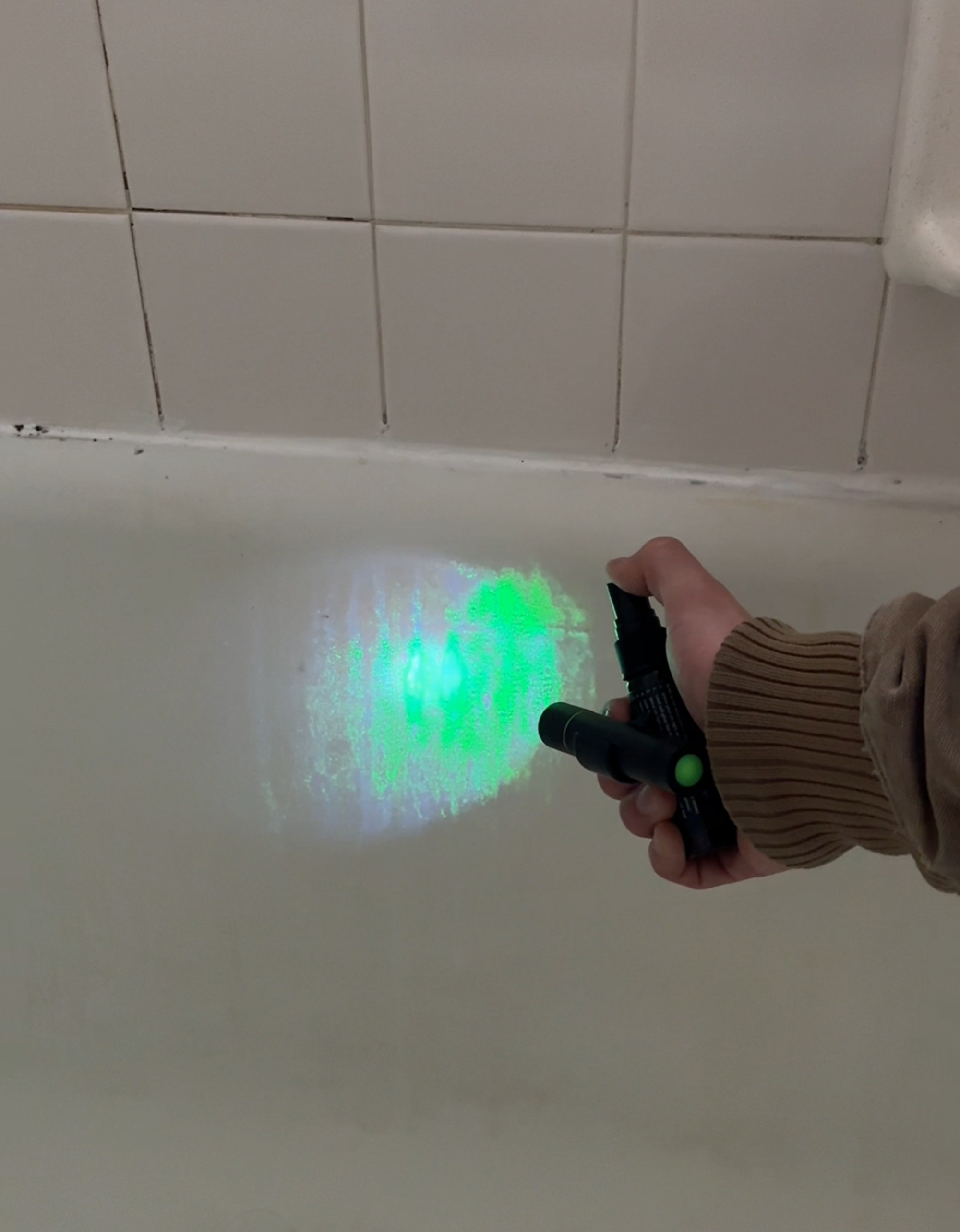 A hand shines a green light from a black flashlight onto a white tiled surface.