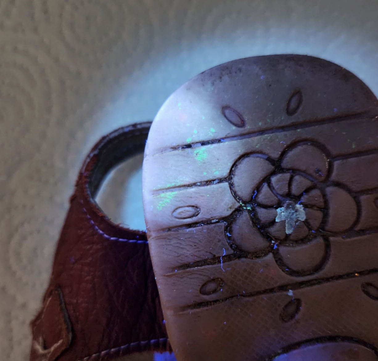 The sole of a shoe with a flower tread under a blacklight, showing glowing green and blue particles.
