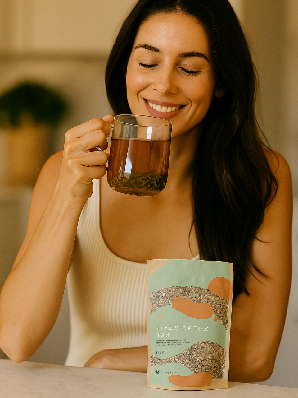 Woman smiling, holding a glass of tea, with a liver detox tea package on the table.