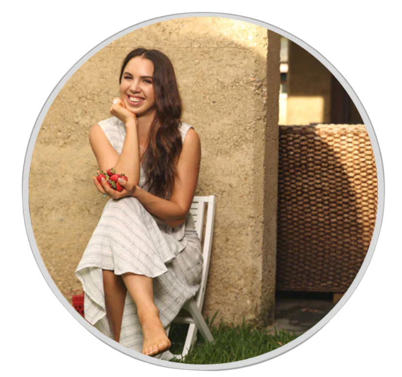 A smiling woman in a striped dress sits on a chair outdoors holding a handful of strawberries.