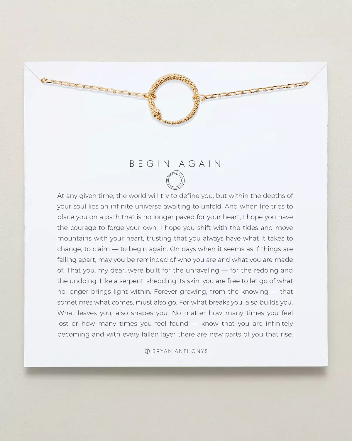 A gold paperclip chain necklace with a textured circular gold pendant is displayed on a white card. The card features the title 'BEGIN AGAIN.