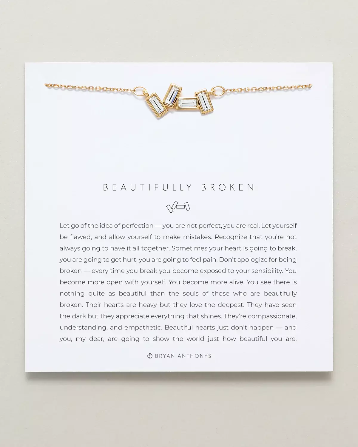 A gold chain necklace with a charm featuring three rectangular, clear baguette stones set closely together, is displayed on a white card. The card features the title 'BEAUTIFULLY BROKEN'.