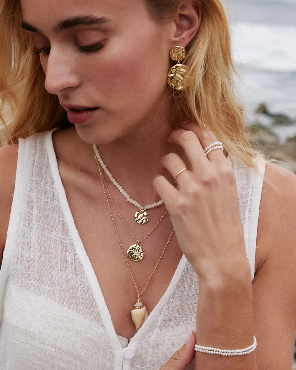 A close-up portrait of a blonde woman wearing a white, textured, sleeveless top outdoors near rocks. She is wearing stacked gold double-coin earrings, and several layered necklaces: one beaded with white stones, two gold chains with coin/sand dollar pendants, and one with a large seashell and horn pendant. She is also wearing layered white beaded bracelets and gold rings.