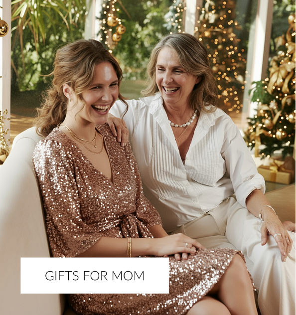 A young woman in a sparkly, rose-gold sequin dress and an older woman in a white collared shirt sit together on a white couch, laughing warmly. Both women are wearing delicate jewelry. The background is brightly lit and decorated for the holidays with golden Christmas lights and ornaments.A white overlay text at the bottom reads, 'GIFTS FOR MOM.'