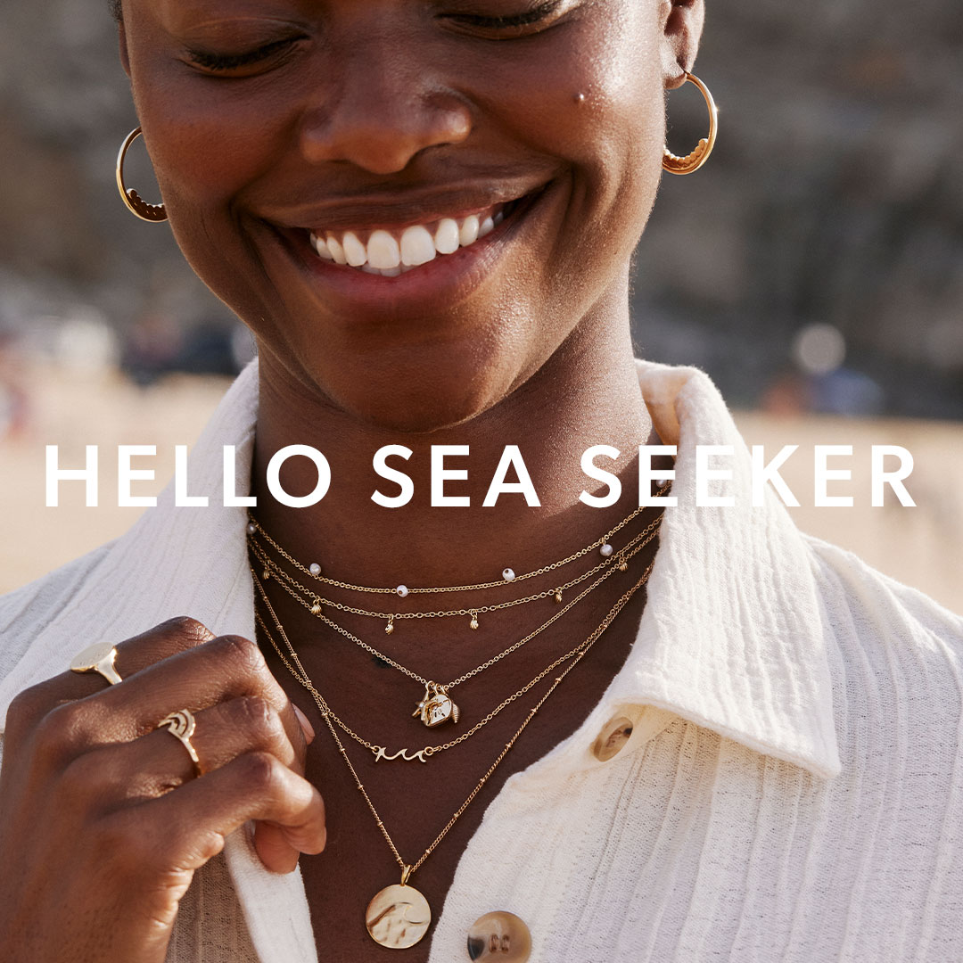 A close-up portrait of a woman wearing a white textured top on a beach. She is smiling and wearing multiple layered gold necklaces, including one with small pearl drops, one shaped like a wave, one with a shell charm, and one with a large coin pendant. She is also wearing gold hoop earrings and several rings. The text overlay reads: 'HELLO SEA SEEKER'.