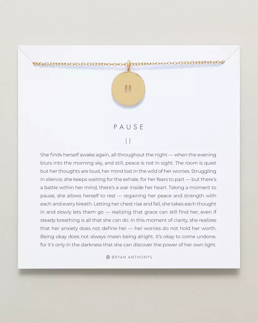A gold chain necklace with a small round gold pendant engraved with a vertical pause symbol, displayed on a white card. The card features the title 'PAUSE.
