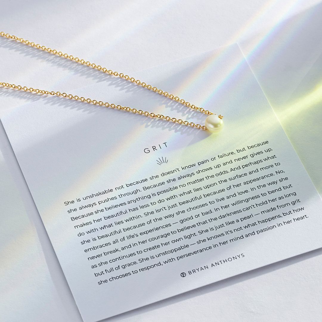 A close-up of a delicate gold chain necklace with a single white pearl pendant, resting on a white card. The card features the title 'GRIT' and a lengthy, inspirational quote describing a strong woman who is 'just like a pearl — made from grit but full of grace.