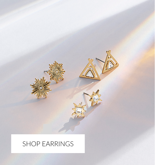 Three pairs of small gold stud earrings arranged on a white surface with bright sunlight and rainbow glare. The styles include a sunburst design, a triangle/teepee design with a clear central stone, and a star shape with a square center stone. White overlay text at the bottom reads 'SHOP EARRINGS.'