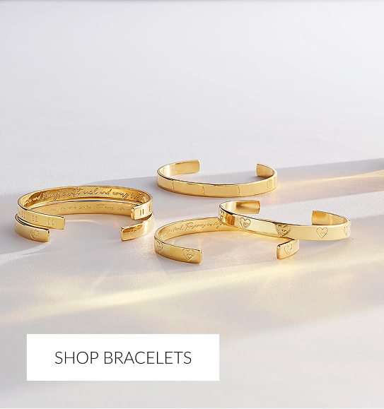 Four gold open cuff bracelets scattered on a white surface, catching direct light. The bracelets are engraved with text and have small heart-shaped details or simple indentations. White overlay text at the bottom reads 'SHOP BRACELETS.'