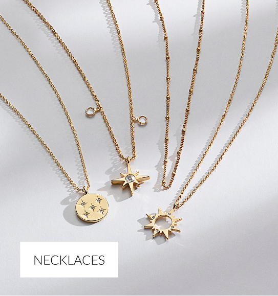 An overhead arrangement of four delicate gold necklaces with celestial-themed pendants on a white surface. The pendants include a round disc with embedded stars, a starburst/compass rose with a center stone, a beaded chain, and a sunburst design. White overlay text at the bottom reads 'NECKLACES.'