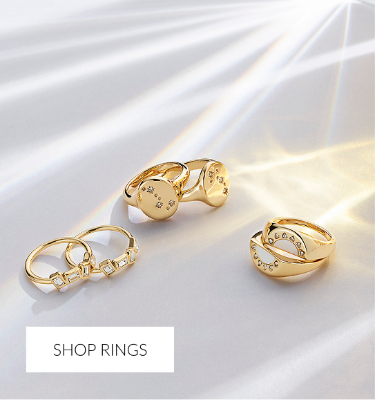 Four gold open cuff bracelets scattered on a white surface, catching direct light. The bracelets are engraved with text and have small heart-shaped details or simple indentations. White overlay text at the bottom reads 'SHOP BRACELETS.'