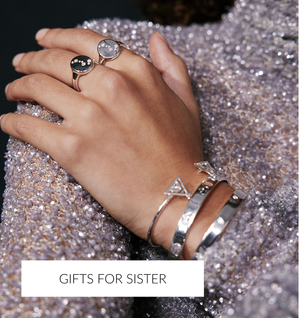 A close-up of a hand resting on a sparkling, lilac sequin garment, showcasing several pieces of jewelry. The hand wears two rose-gold rings (one black, one gray with stars) and two silver bangles/bracelets with triangle accents. A white overlay text at the bottom reads, 'GIFTS FOR SISTER.
