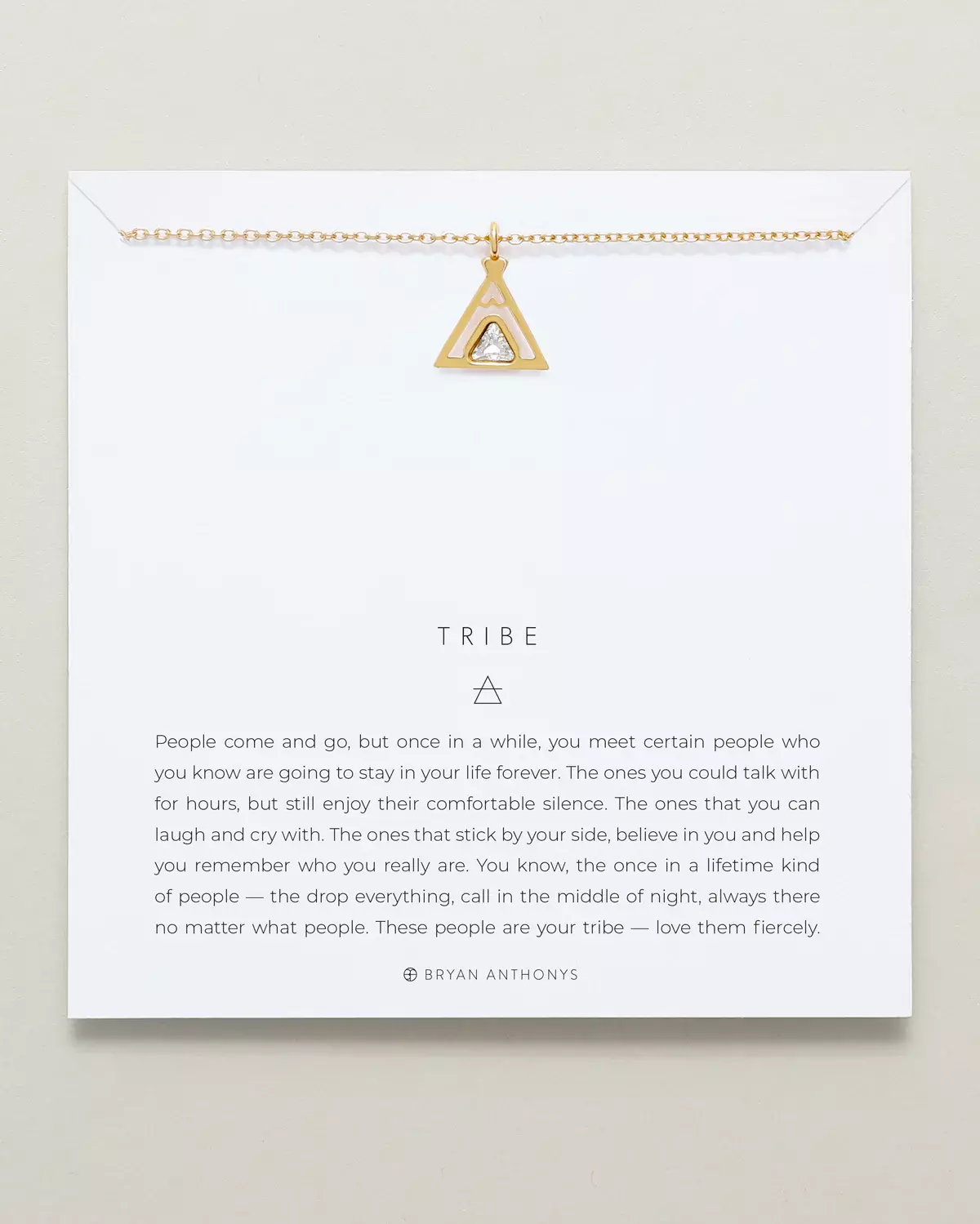 A gold chain necklace with a triangular pendant that outlines a teepee shape and holds a clear center stone, is displayed on a white card. The card features the title 'TRIBE'.