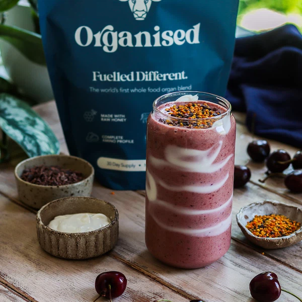 Pink smoothie in a swirl-patterned glass with toppings, surrounded by bowls and a blue package.