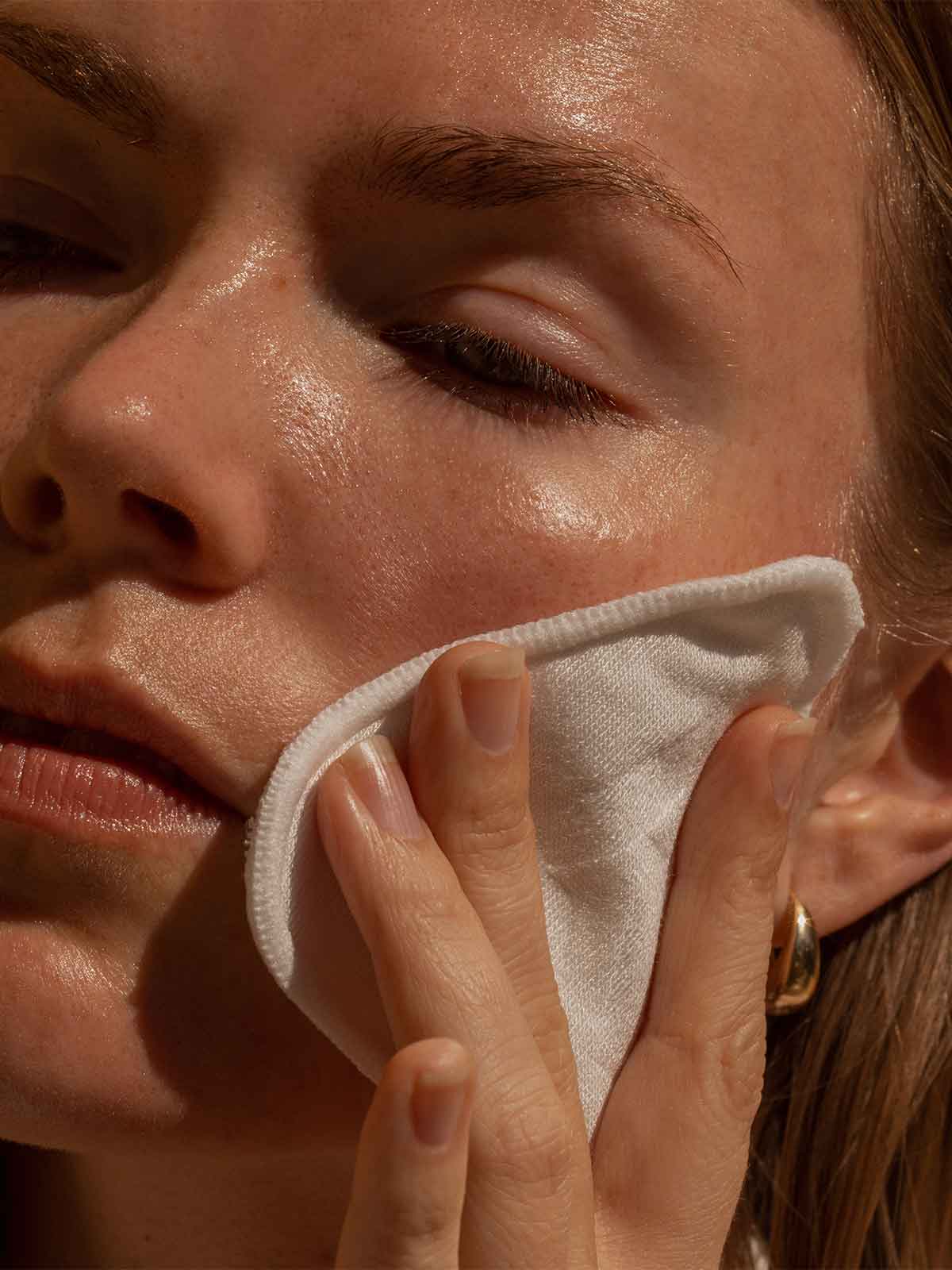A close-up of a person with glowing skin wiping their cheek with a white pad.