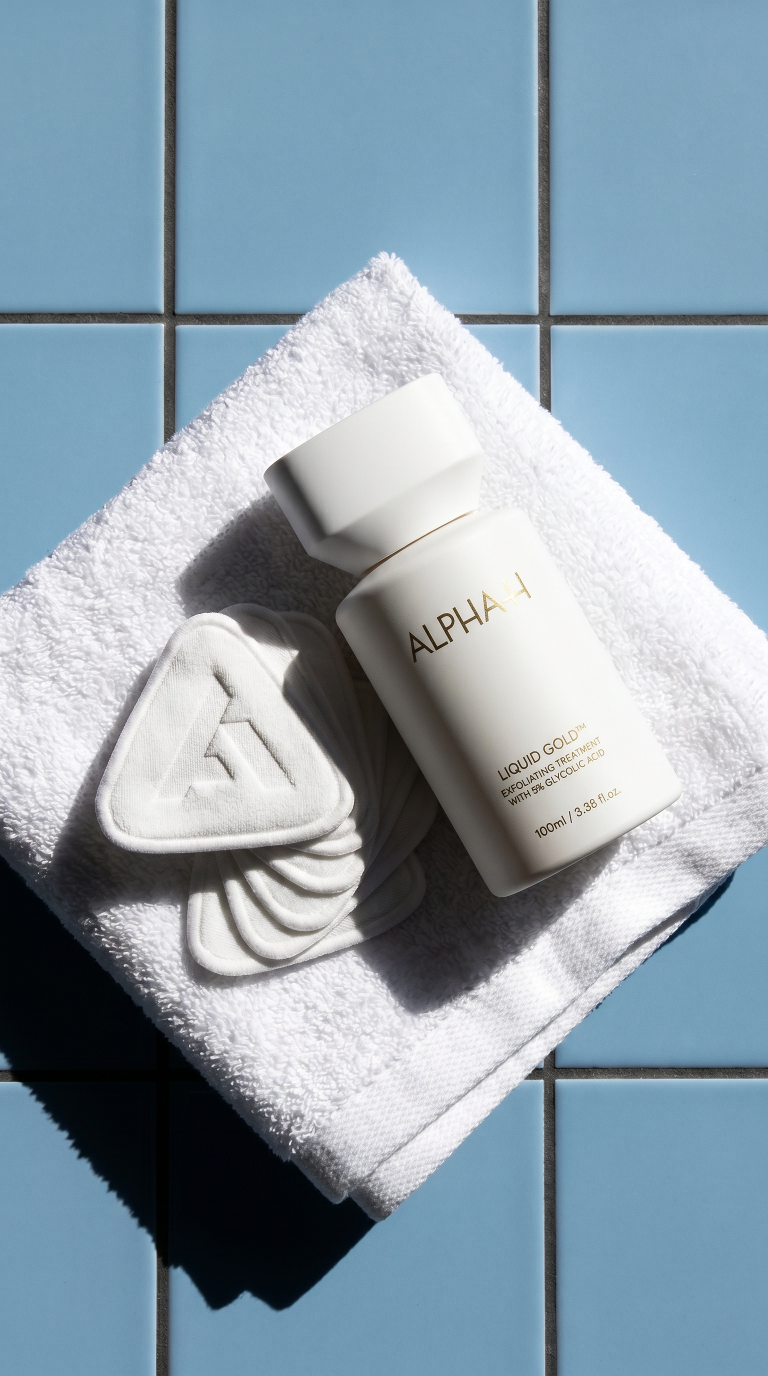 An Alpha-H Liquid Gold bottle and triangular pads on a white towel against blue tiles.