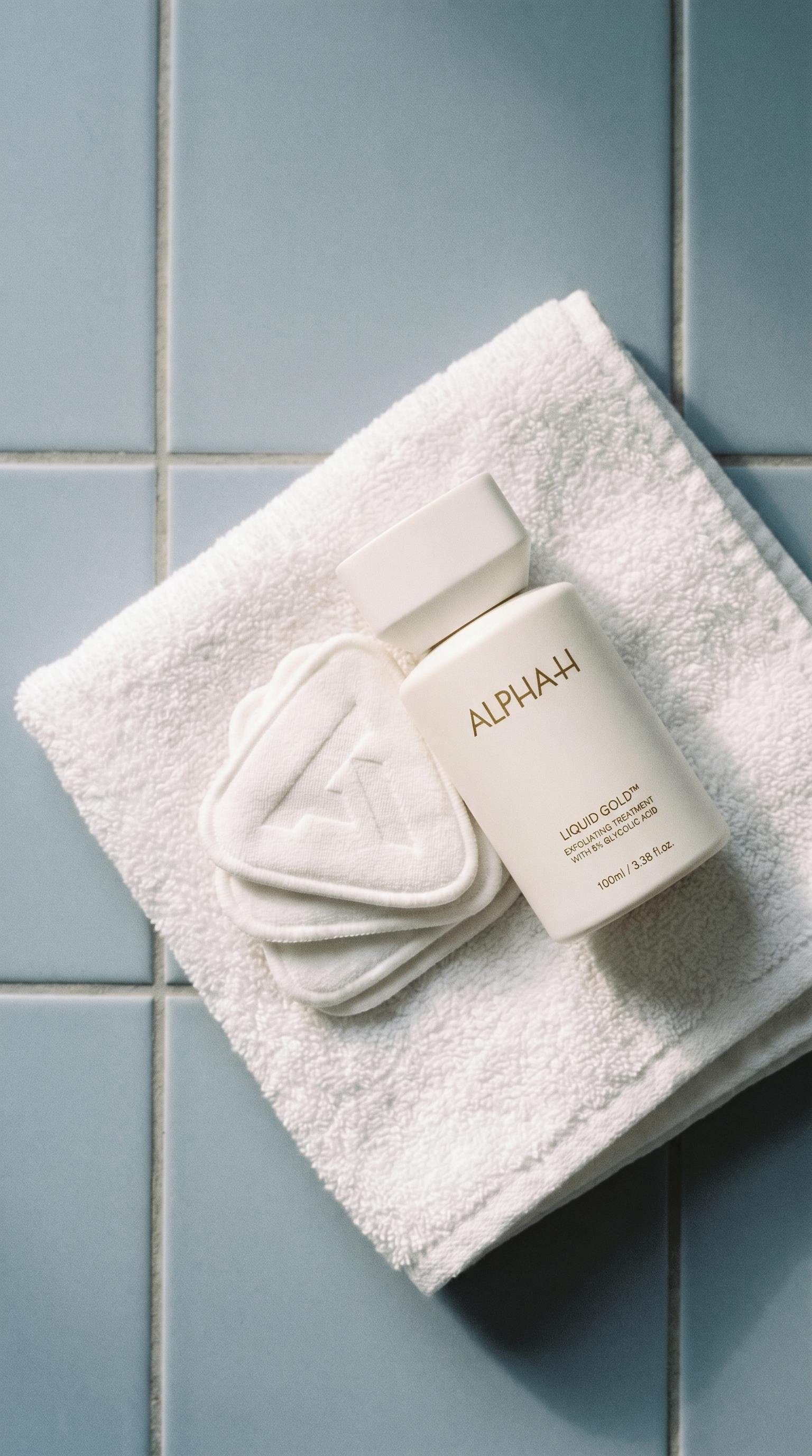 A white bottle of Alpha-H Liquid Gold with triangular pads resting on a white towel on blue tiles.