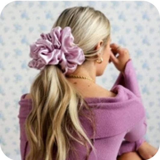 Woman with blonde hair in a ponytail, wearing a large pink scrunchie and purple sweater.