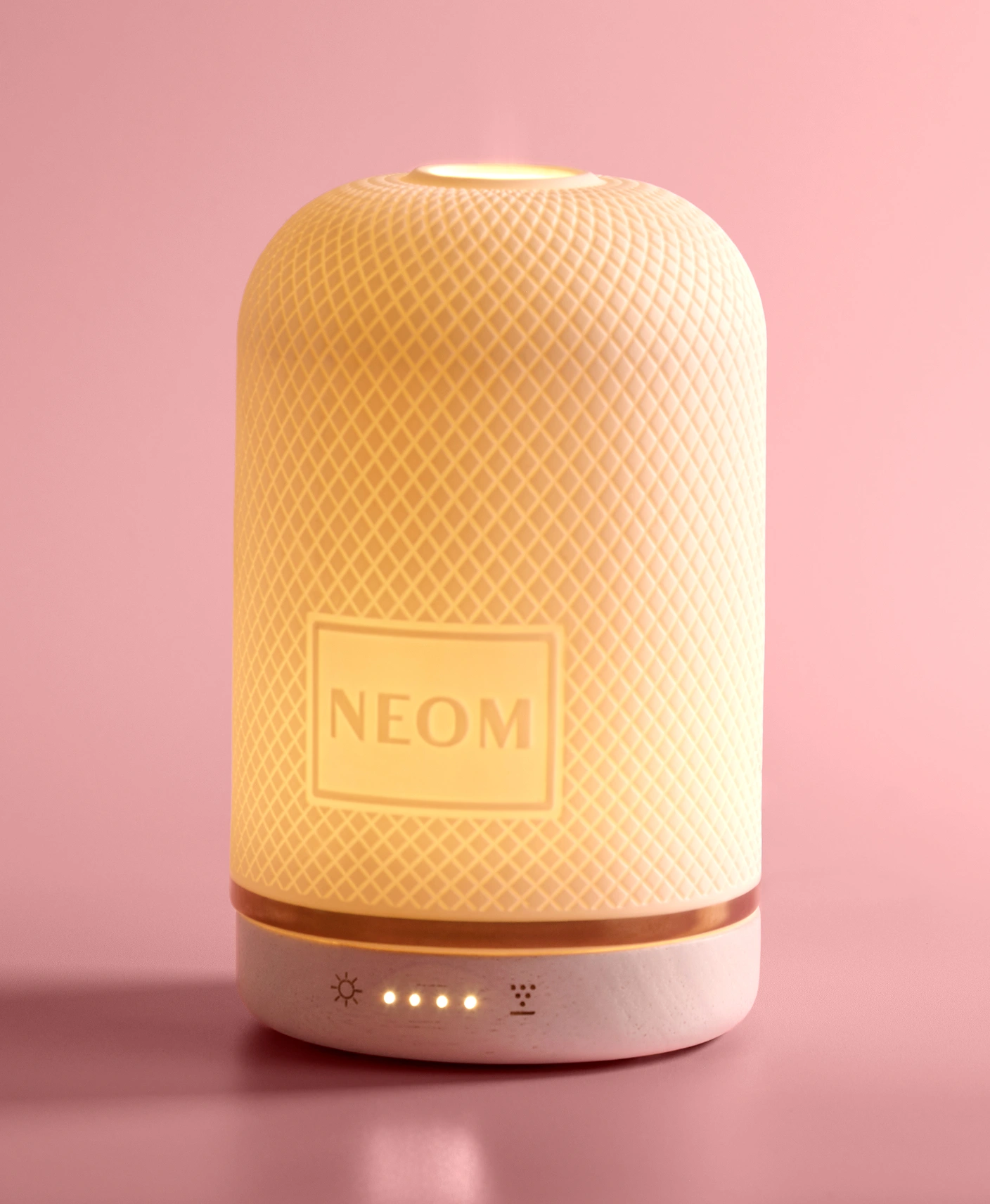 A white, illuminated Neom aroma diffuser with a diamond pattern, set against a pink background.