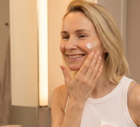 A smiling blonde woman applying a dab of white cream to her cheek while looking in a mirror.