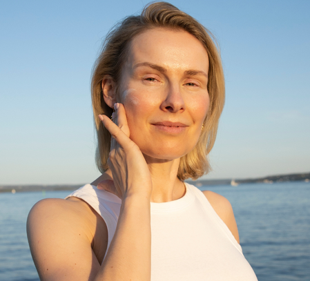 A blonde woman in a white top touches her cheek while standing in front of a body of water.