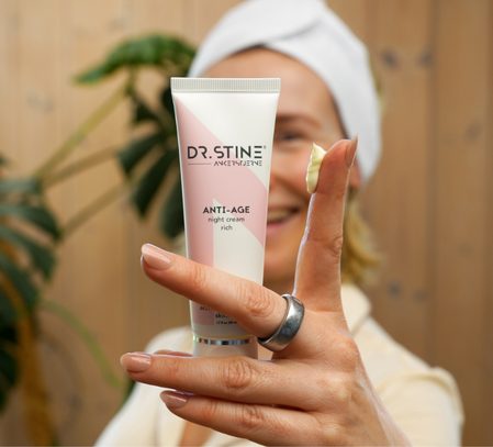 A smiling woman holds a tube of Dr. Stine night cream with a dab of it on her finger.
