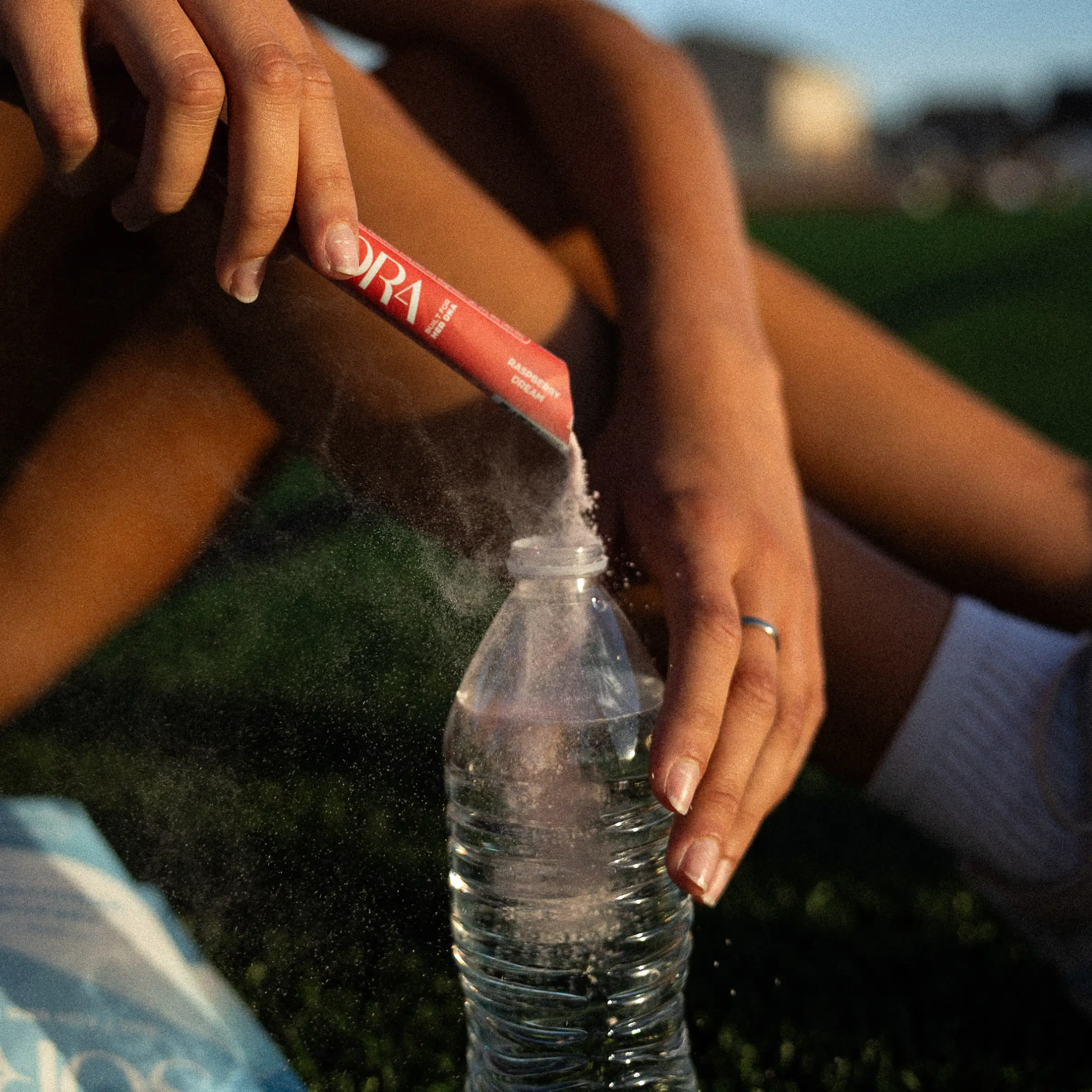 A person pours a red packet of powder into a plastic water bottle while sitting on grass.