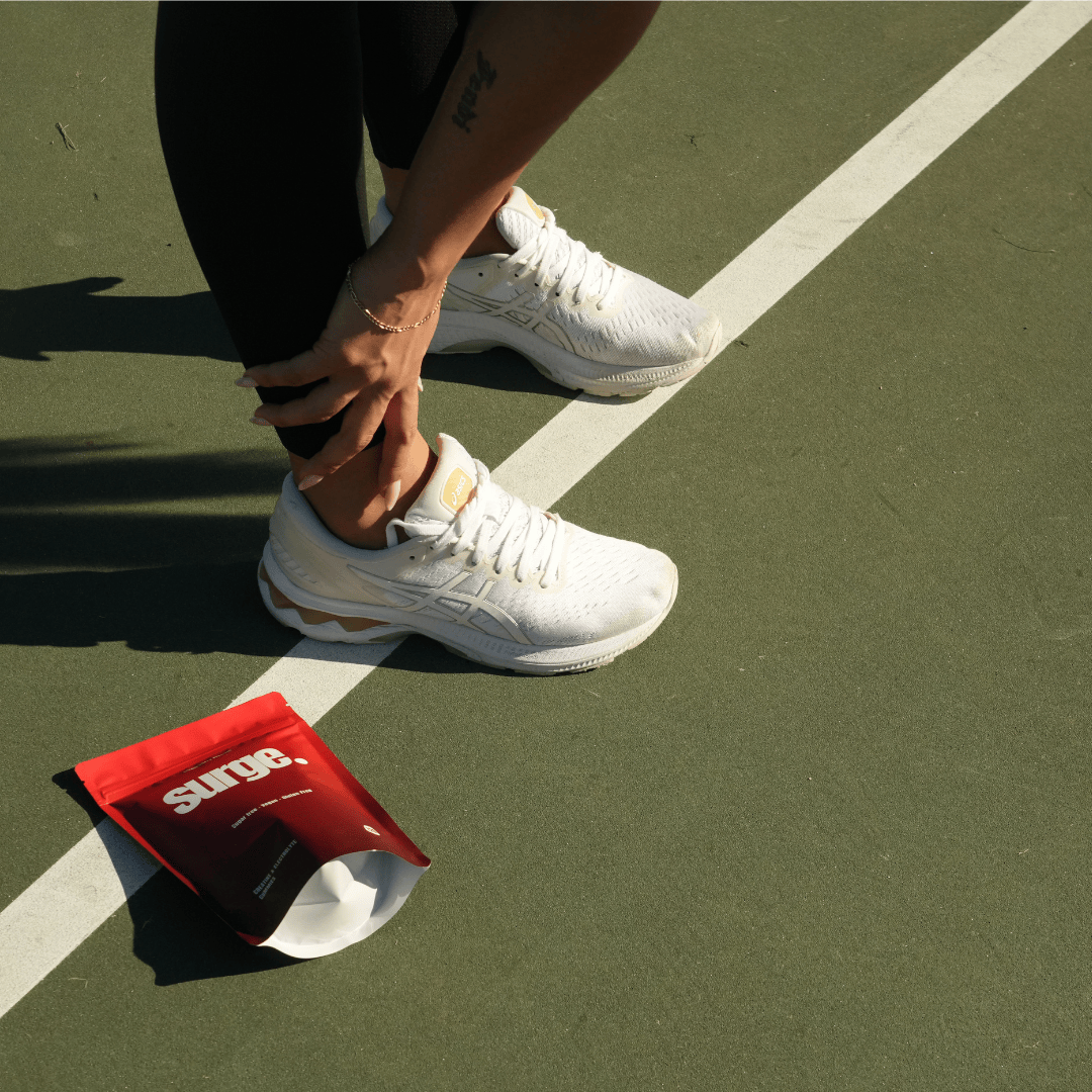Person in white sneakers adjusting ankle on a sports court, with a red pouch nearby.