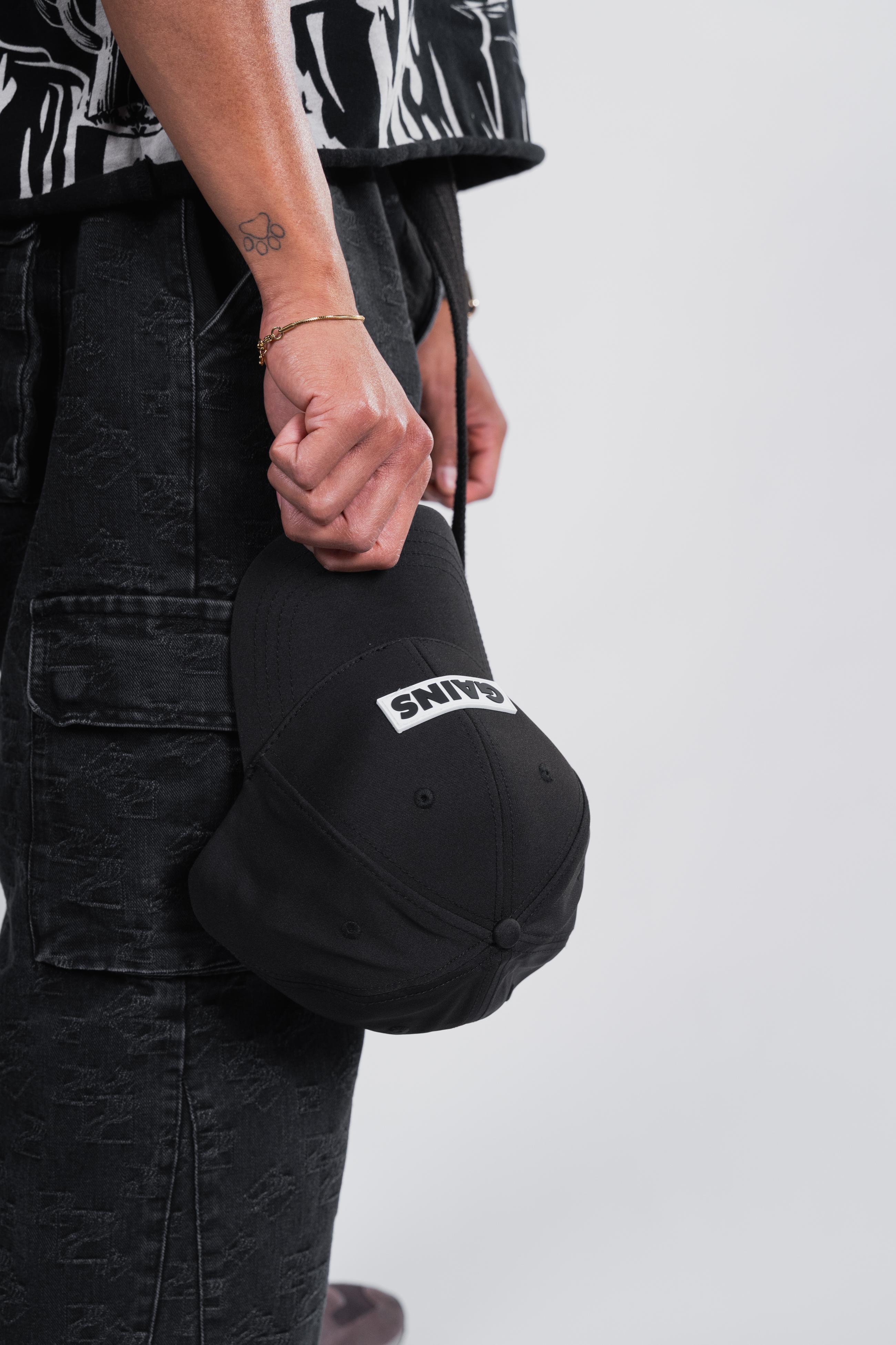 Person holding a black cap with 'GAINS' text, wearing dark jeans.