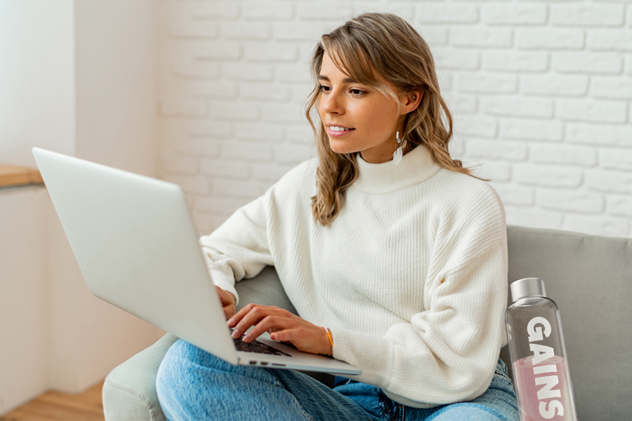 Woman in white sweater using a laptop with a bottle labeled 'GAINS' beside her.