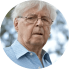 A circular headshot of an older man with white hair and glasses, wearing a blue striped shirt.
