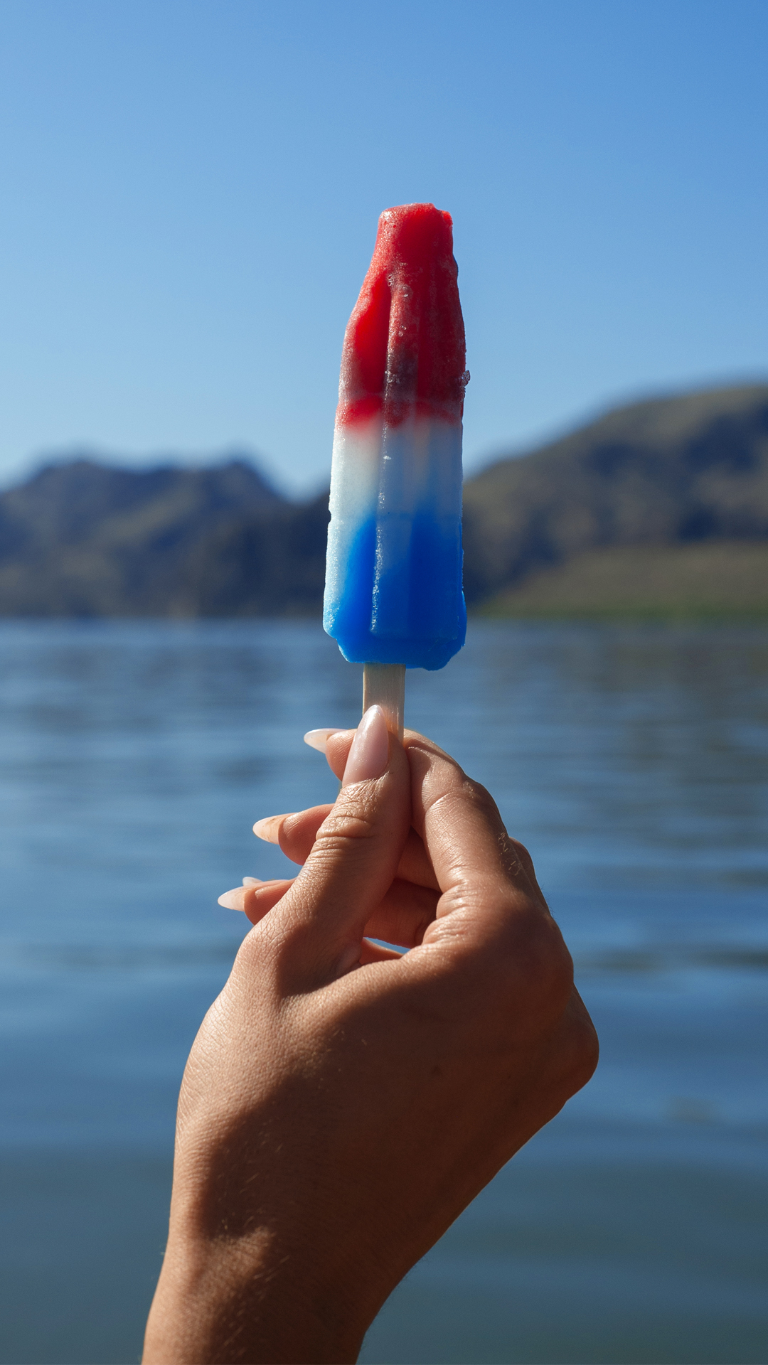 Hand holding a red, white, and blue popsicle by a lake.