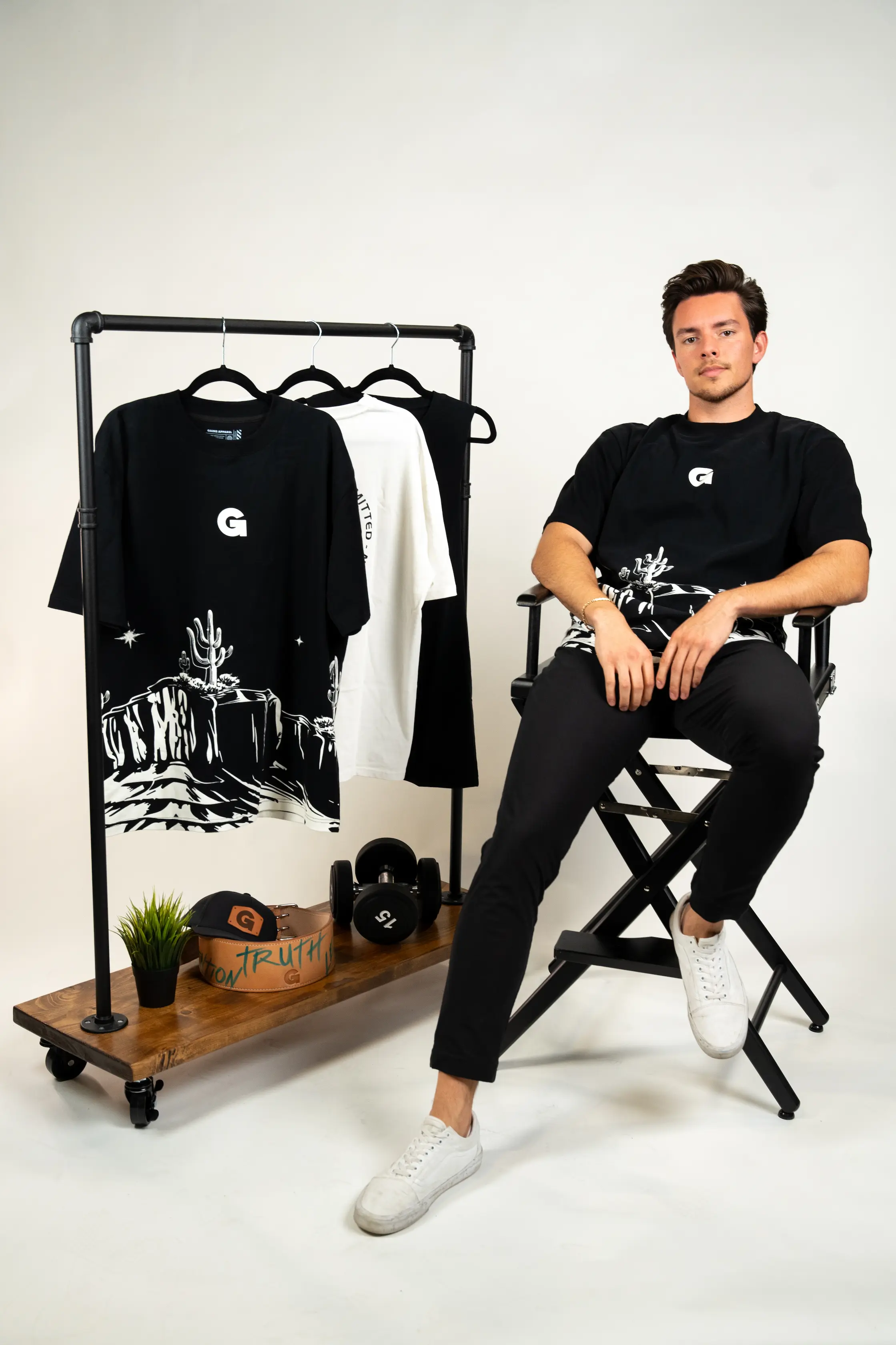 Man sitting beside clothing rack with t-shirts, cap, and dumbbells.