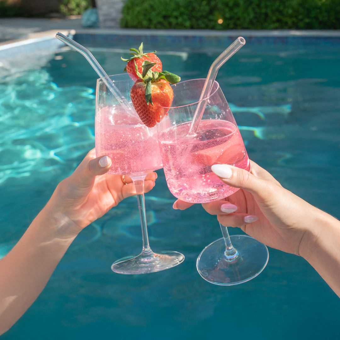 Two hands clinking glasses of pink drinks by a pool.