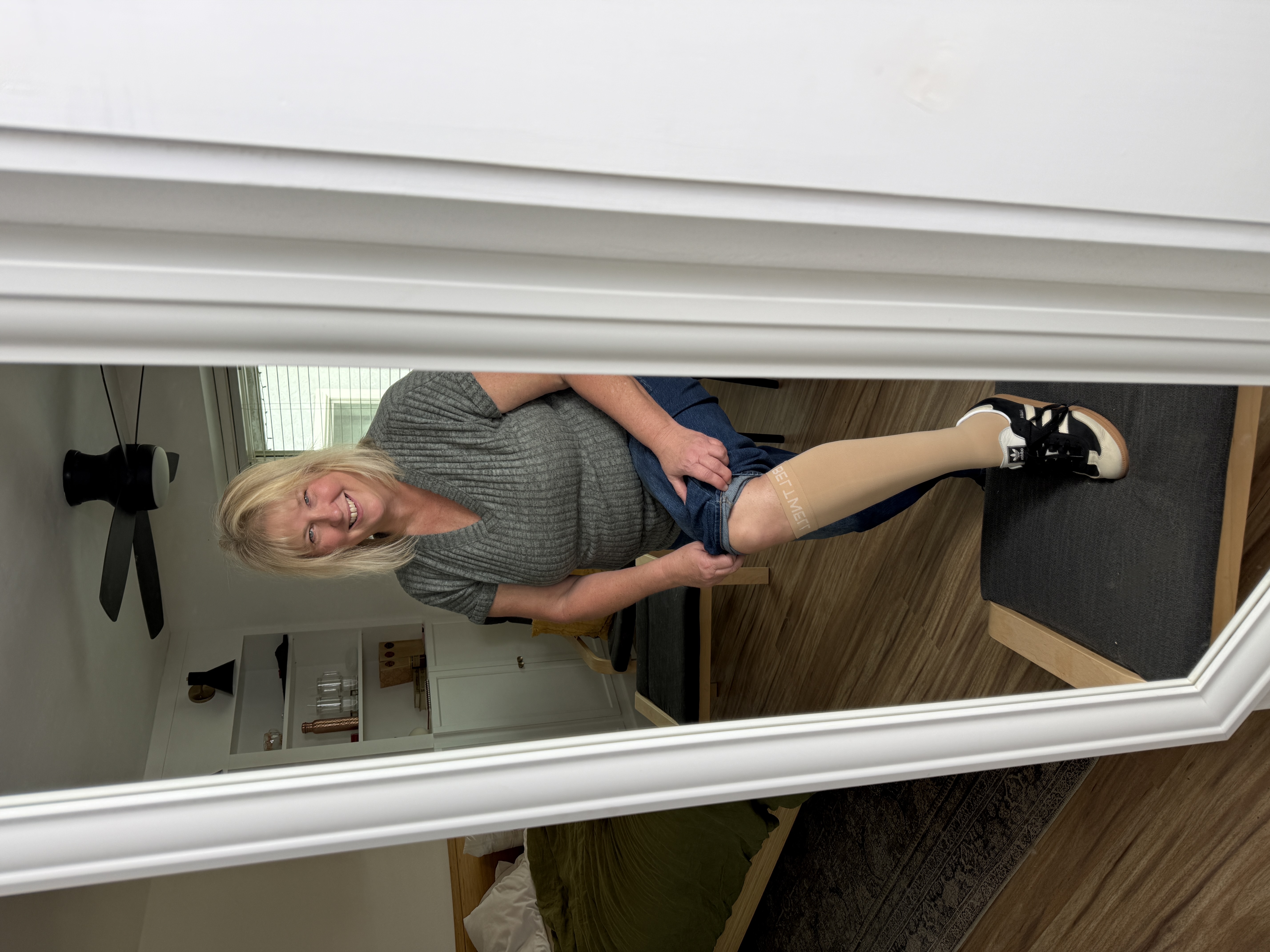 A smiling woman looks in a mirror while sitting and pulling up a tan compression sock.
