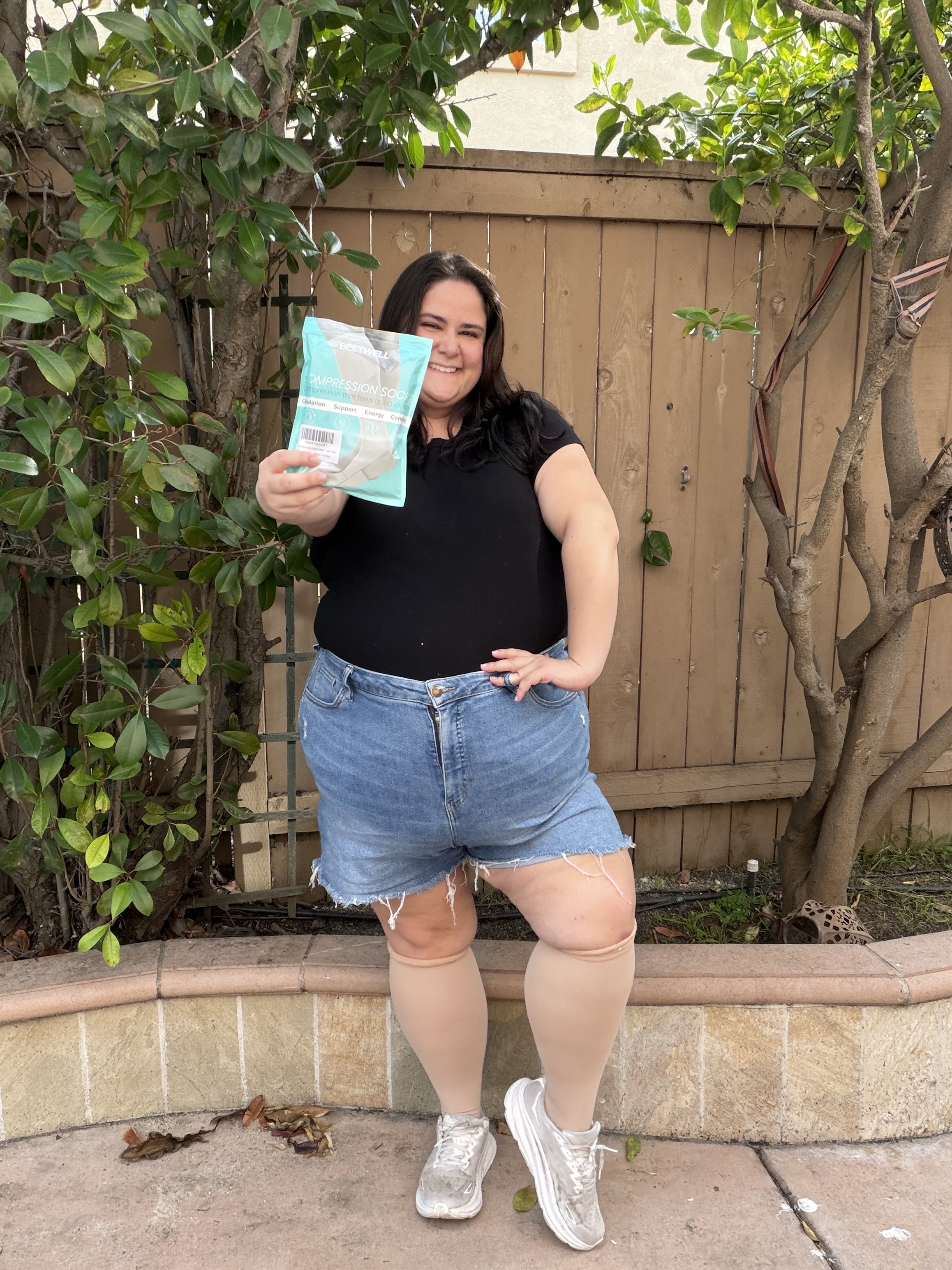 A smiling woman stands outside holding a package of compression socks while wearing a pair.